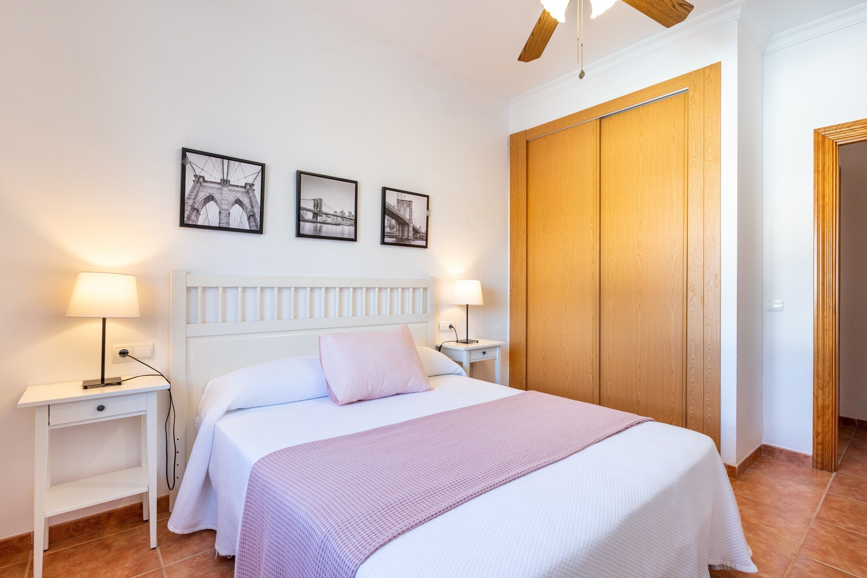 Een slaapkamer met een bed, twee nachtkastjes, een nachtkastje, een houten garderobe en drie ingelijste foto's aan de muur.