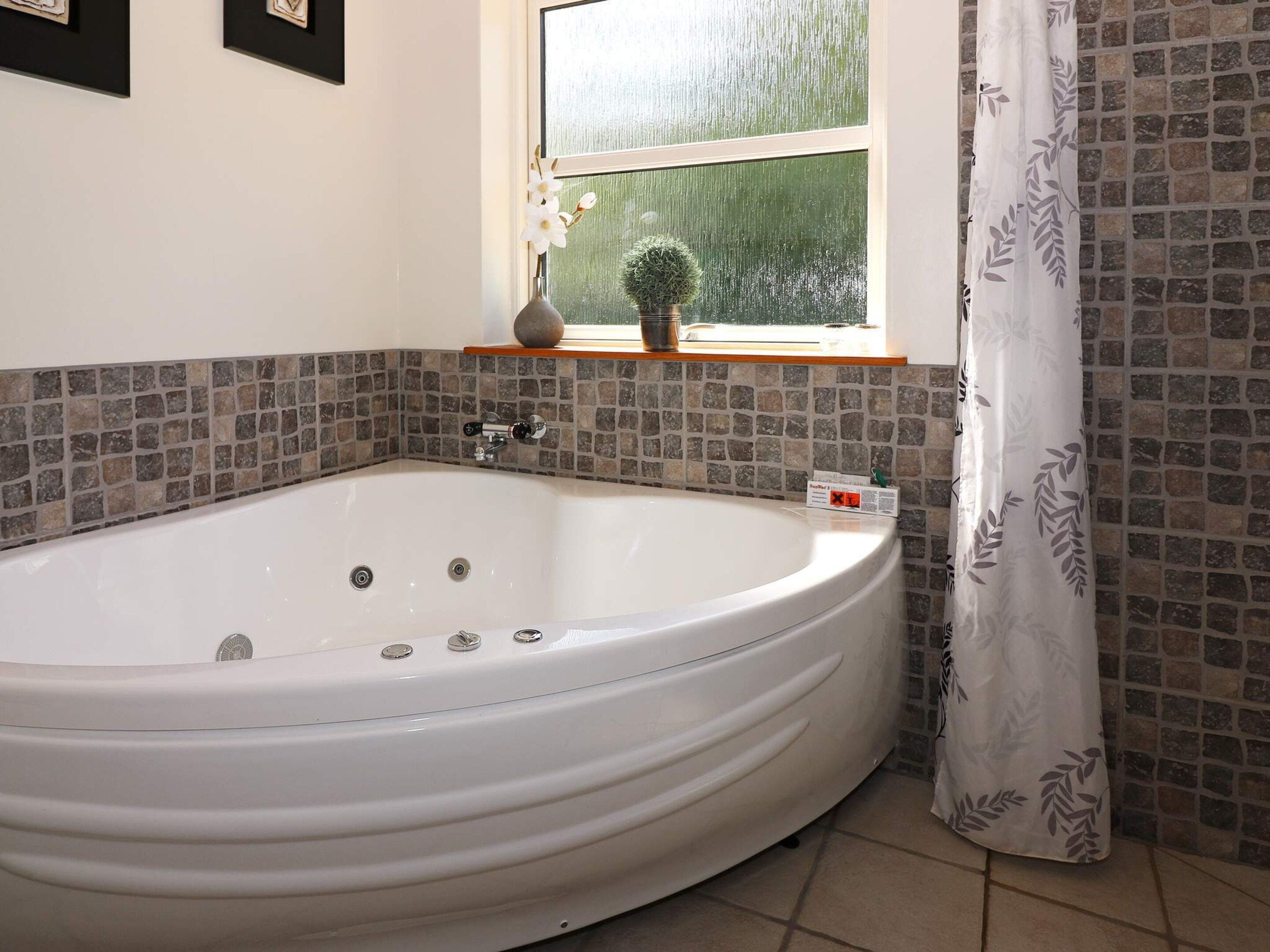A bathroom with a large white bathtub, a window with a view of greenery, a small shelf with a plant, and a tiled backsplash.