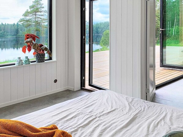 A bedroom with a large window, a bed, and a view of a lake and forest.