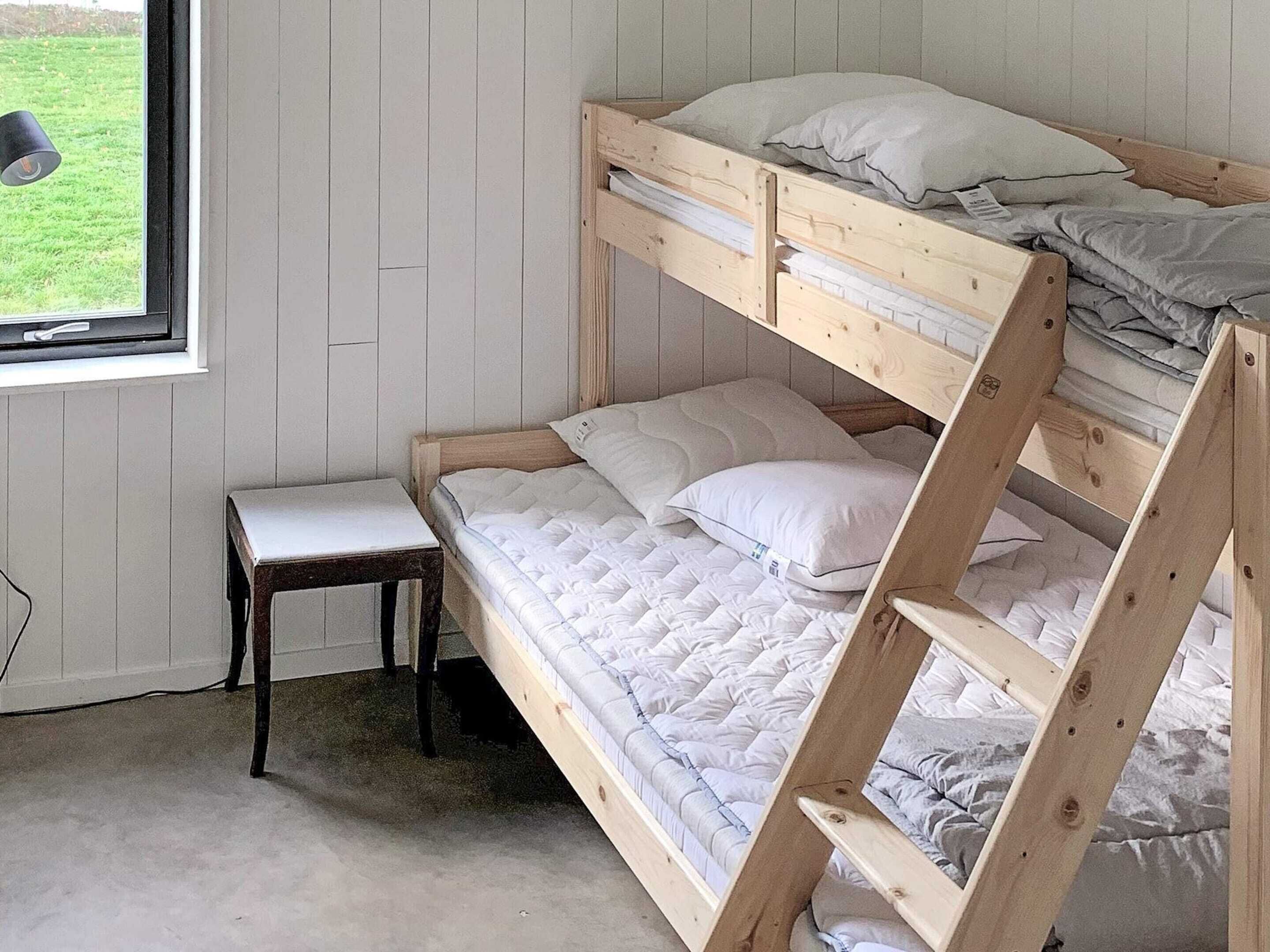 A bunk bed with a wooden frame and white bedding, a small bedside table, and a window with a view of a grassy area.