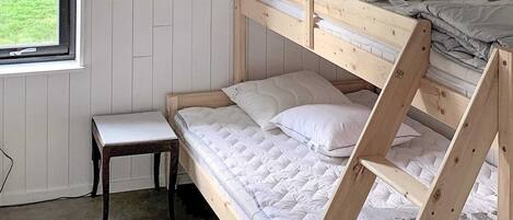 A bunk bed with a wooden frame and white bedding, a small bedside table, and a window with a view of a grassy area.