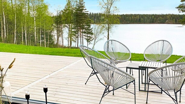 Una terrazza in legno con sedie in vimini bianco e un tavolo che si affaccia su un lago, circondata da alberi.