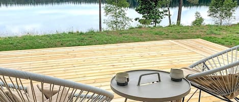 A wooden deck with two chairs and a table, overlooking a calm lake with trees in the background.