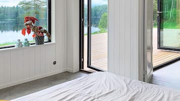 Un dormitorio con una cama, un ventanal con vistas a un lago y un bosque, y una terraza de madera afuera.