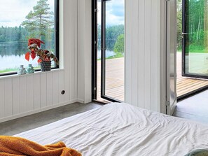 A bedroom with a bed, a window with a view of a lake and forest, and a wooden deck outside.