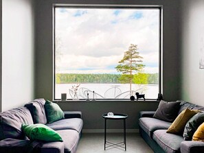 A room with two grey sofas, a small round table, and a large window overlooking a lake with trees and a clear sky.