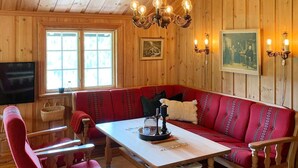 Hyggelig hytteinteriør med træpaneler, en sofa, stole, et bord, en lysekrone og kunst på væggene.