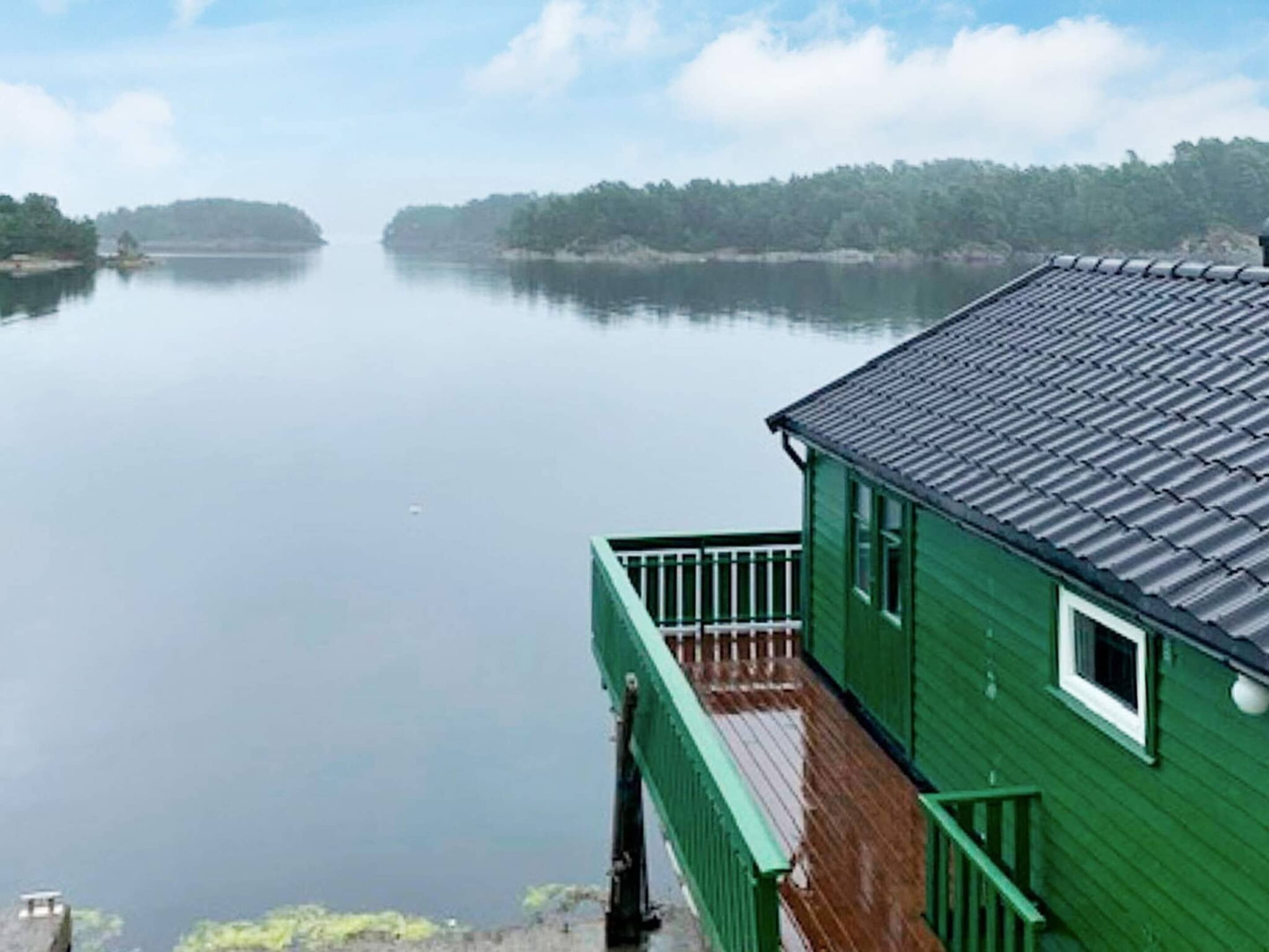 Un bâtiment écologique doté d’une terrasse en bois et d’un toit en métal, offrant une vue sur un lac paisible et une terre lointaine.