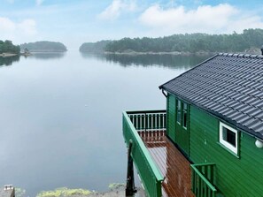 A green building with a wooden deck and a metal roof, overlooking a calm lake with distant trees. - 6 Person Holiday Home in Lindas (Alver)