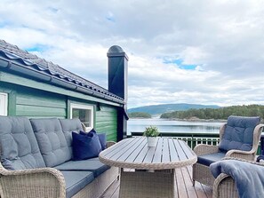 A deck with a sofa, chairs, and a table, overlooking a body of water and hills. - 6 Person Holiday Home in Lindas (Alver)