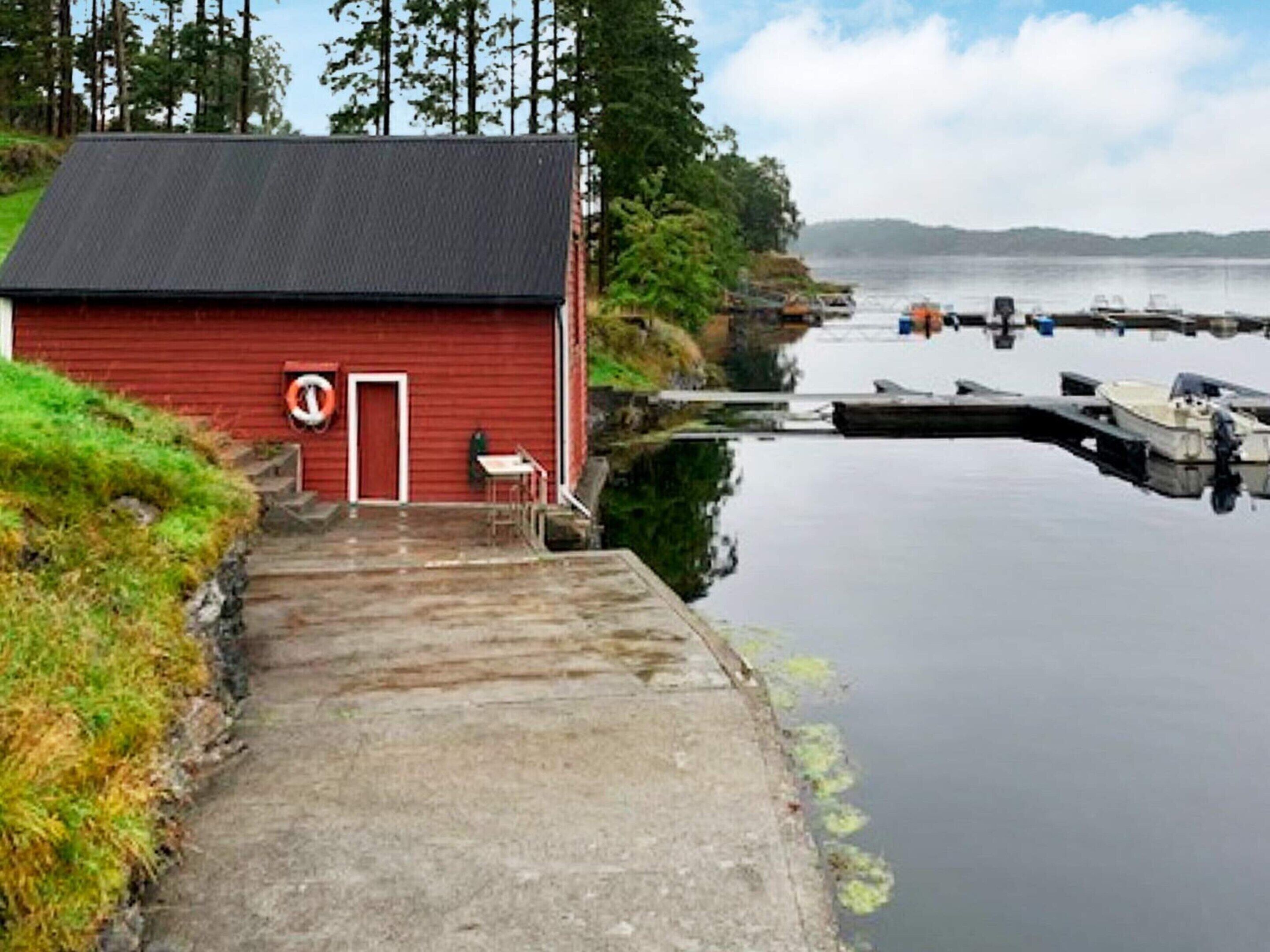 Un bâtiment rouge avec une porte et une petite table à l’extérieur, près d’une étendue d’eau calme où se trouvent des bateaux.