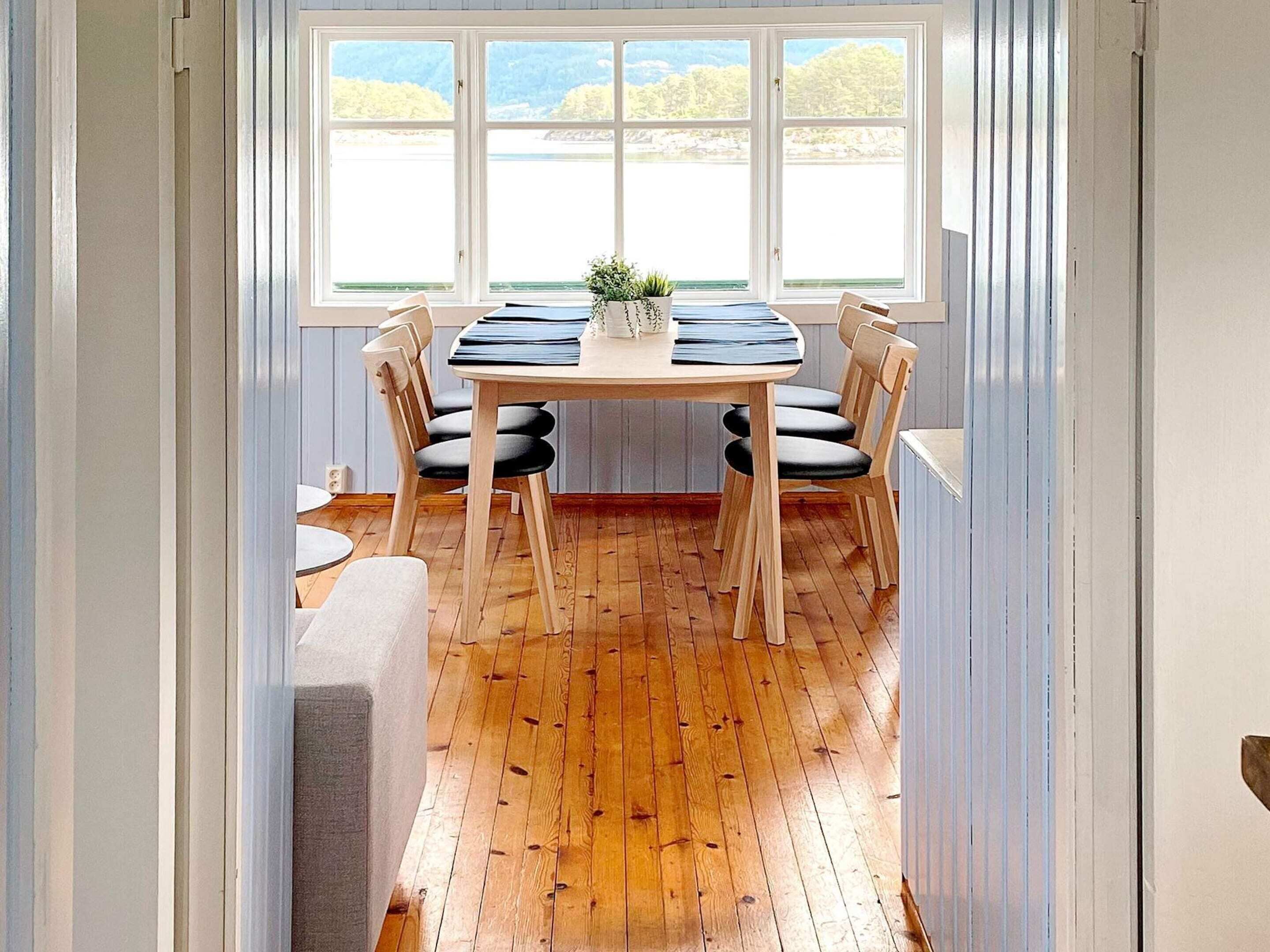 A dining area with a wooden table, four chairs, a small plant, and a view through large windows.