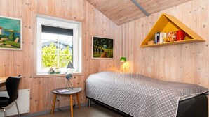 A bedroom with a bed, a desk, a chair, a small table, a window with a view, and a shelf with books and a red mug. - Holiday Home in Rømø (Rømø)