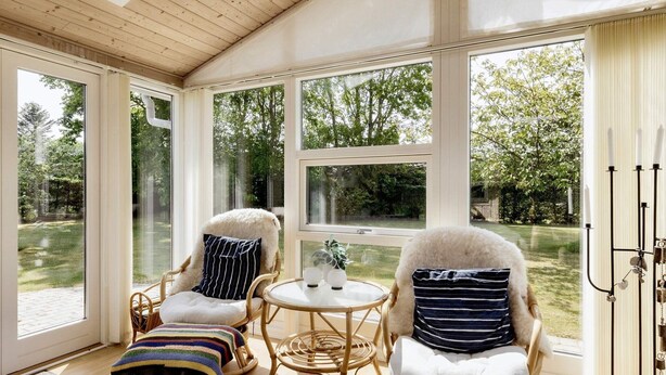 A cozy room with two wicker chairs, a small round table, and a view of a lush garden through large windows.