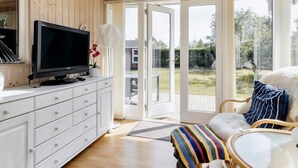 A modern living room with a flat-screen TV on a white cabinet, a sofa with a colorful blanket, and a glass door leading to a garden.