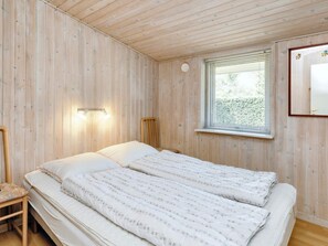 Una habitación con paredes de madera y una cama con ropa blanca. Hay una ventana con persianas y una lámpara fijada a la pared.