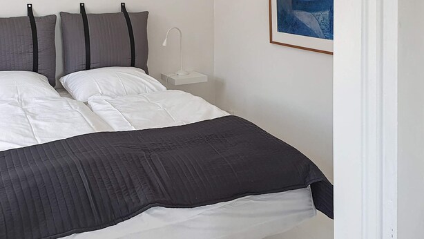 A neatly made bed with white and dark bedding, a bedside table with a lamp, and a framed picture on the wall. - Holiday Home in Snedsted (Snedsted)