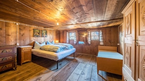 Een slaapkamer in een houten hut met een bed, een stoel, een dressoir en twee ramen.