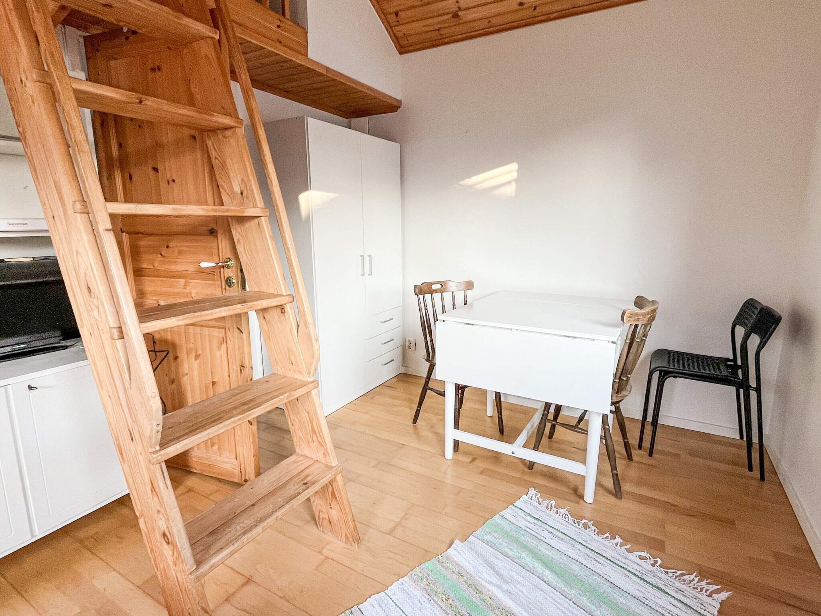 Una habitación con una escalera de madera, armarios blancos, una mesa blanca y dos sillas.