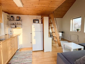 Una cocina funcional con refrigerador, microondas y cafetera, una escalera de madera, y un comedor con mesa y sillas blancas.