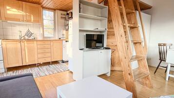 Una cocina moderna con armarios de madera, una encimera blanca y un televisor integrado. Una escalera de madera se apoya contra la pared, y hay una mesa blanca en primer plano.