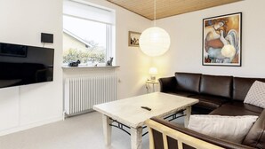 A modern living room with a brown leather sofa, a white table, a mounted TV, a radiator, and a window with a view of greenery. - 6 Person Holiday Home in Aeroskobing (Aeroskobing)