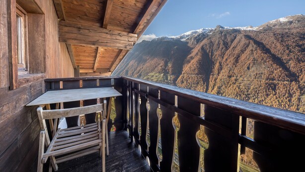 Ein hölzerner Balkon mit Tisch und Stühlen, mit Blick auf eine Berglandschaft mit schneebedeckten Gipfeln.