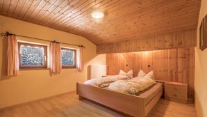 Ein Schlafzimmer mit Holzboden, einem Bett mit gemusterten Bettbezügen, zwei Fenstern mit Vorhängen und einem Nachttisch.
