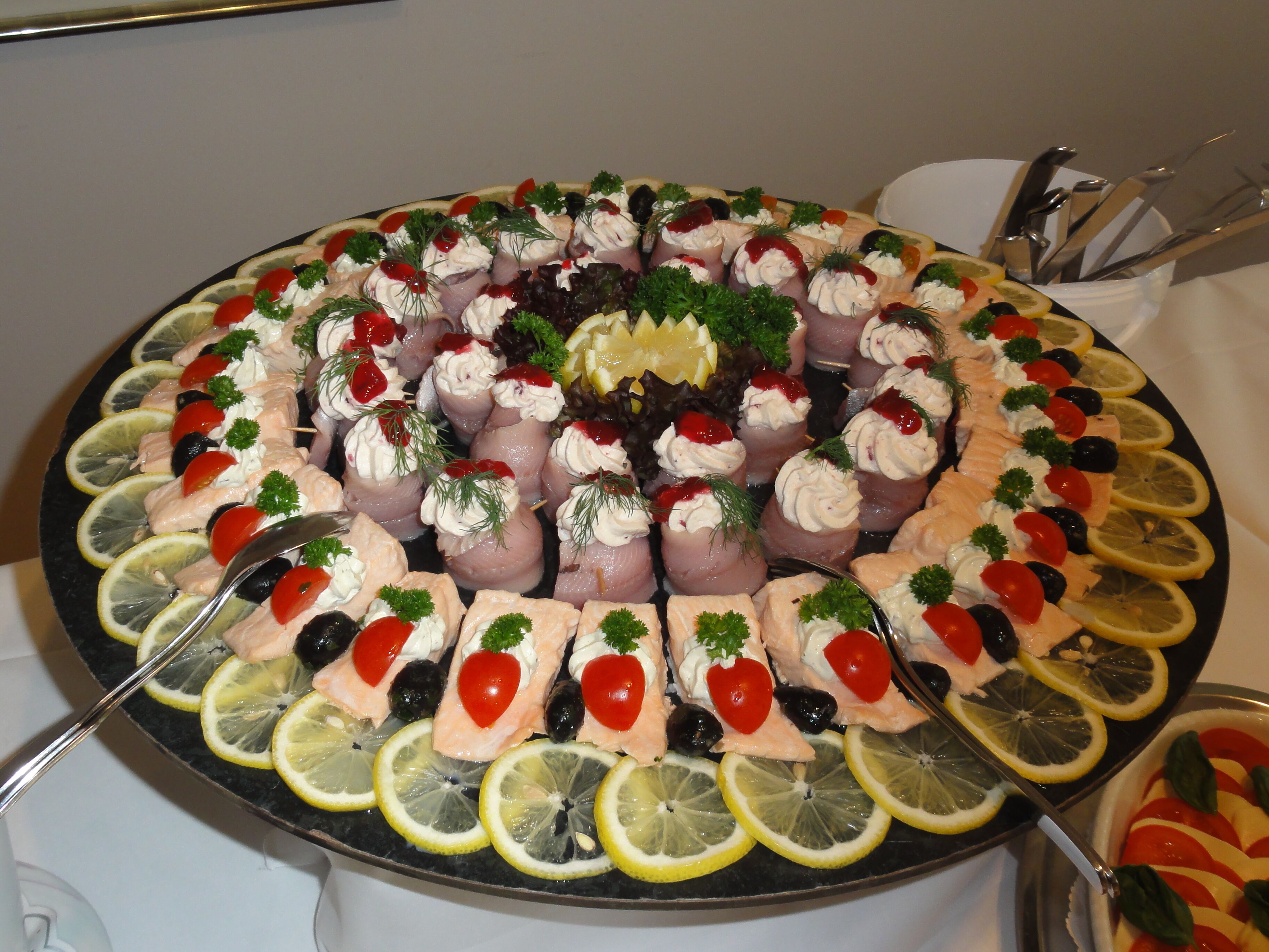 A large platter of food arranged in a circular pattern, featuring slices of salmon, cherry tomatoes, olives, and lemon slices.