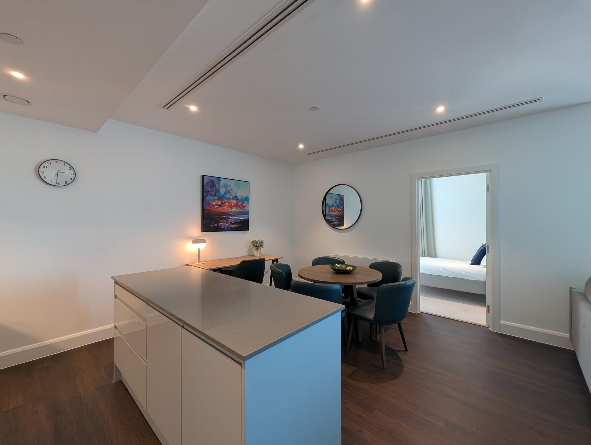 A modern kitchen with a central island, a dining area with blue chairs, a large window with a view, and a clock on the wall.