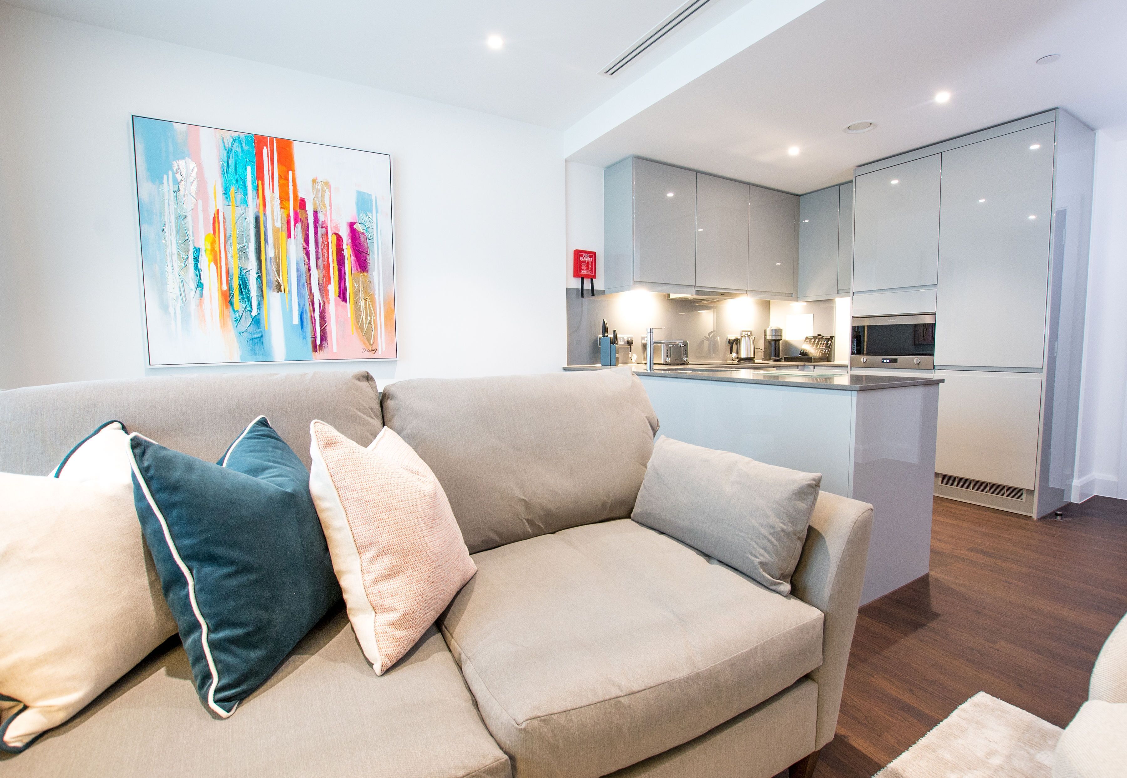 A modern living room with a beige sofa, a colorful abstract painting, and a kitchen with white cabinets and stainless steel appliances.
