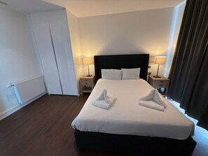 A neatly made bed with two towels folded on it, a black headboard, two bedside lamps, and a window with curtains. - Charles Hope Apartment Canary Wharf (London)