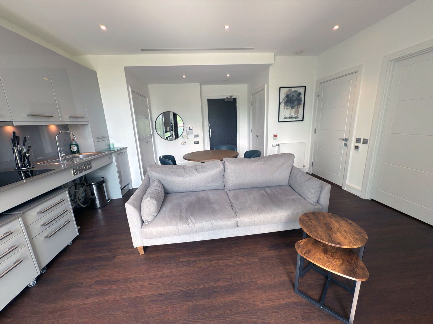 A modern living space with a grey sofa, a wooden stool, a kitchen area with a sink and cabinets, and a dining table with chairs.