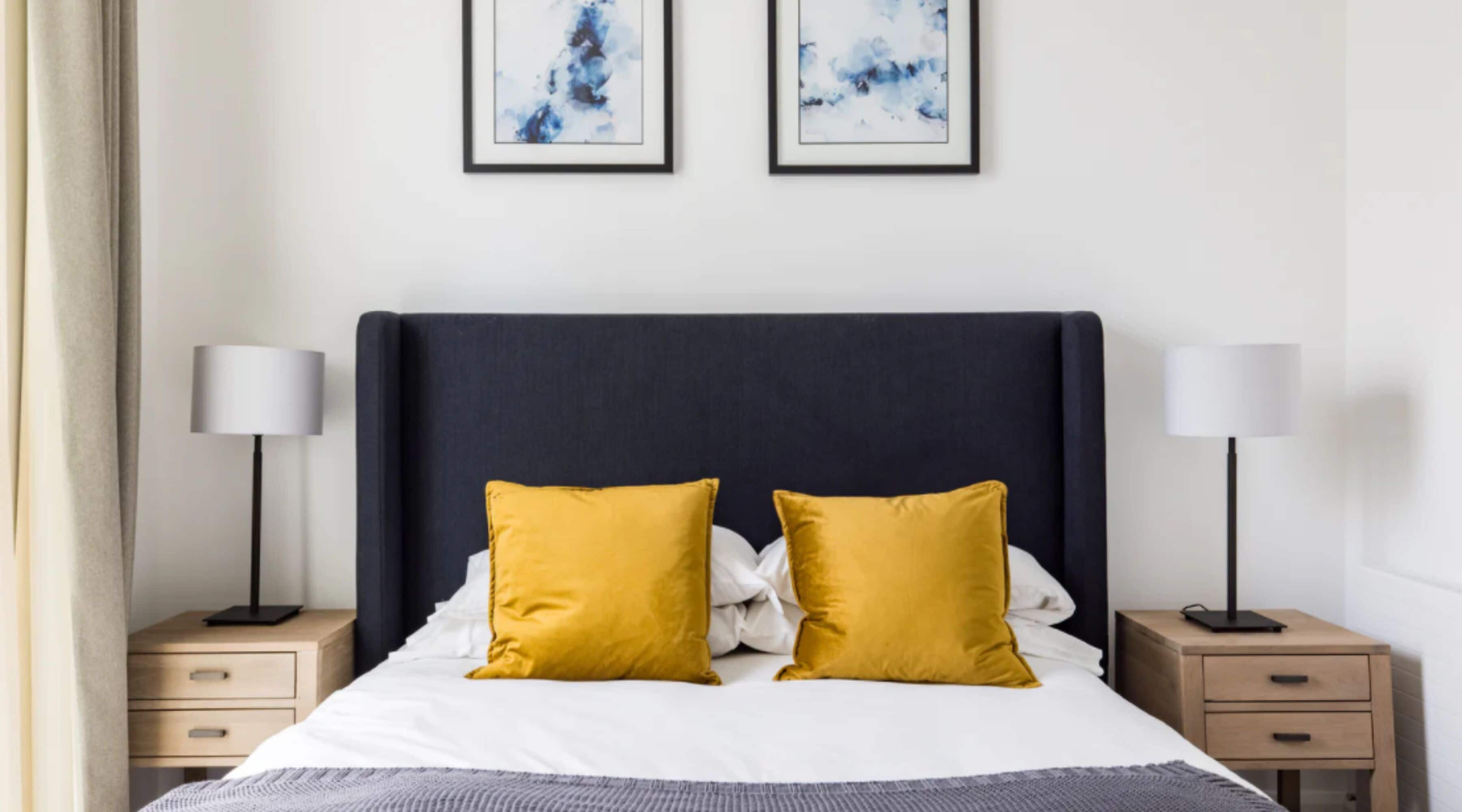 Una cama bien hecha con dos almohadas, dos mesitas de noche con lámparas y dos cuadros abstractos en la pared.