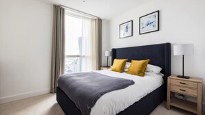 A modern hotel room with a large bed, two bedside tables, a nightstand, and a window with curtains. - Charles Hope Apartment Canary Wharf (London)