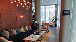 A modern lobby with a dark-colored sofa, a light-colored armchair, and a glass coffee table. There are potted plants and a sign about social distancing. - Charles Hope Apartment Canary Wharf (London)