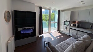 A modern living room with a large flat-screen TV, a sofa, a small table, and a kitchen area with cabinets and appliances. - Charles Hope Apartment Canary Wharf (London)