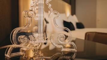 Une lampe en verre transparent au design original, placée sur une table dans une chambre d’hôtel, avec un lit et une table de chevet en arrière-plan.