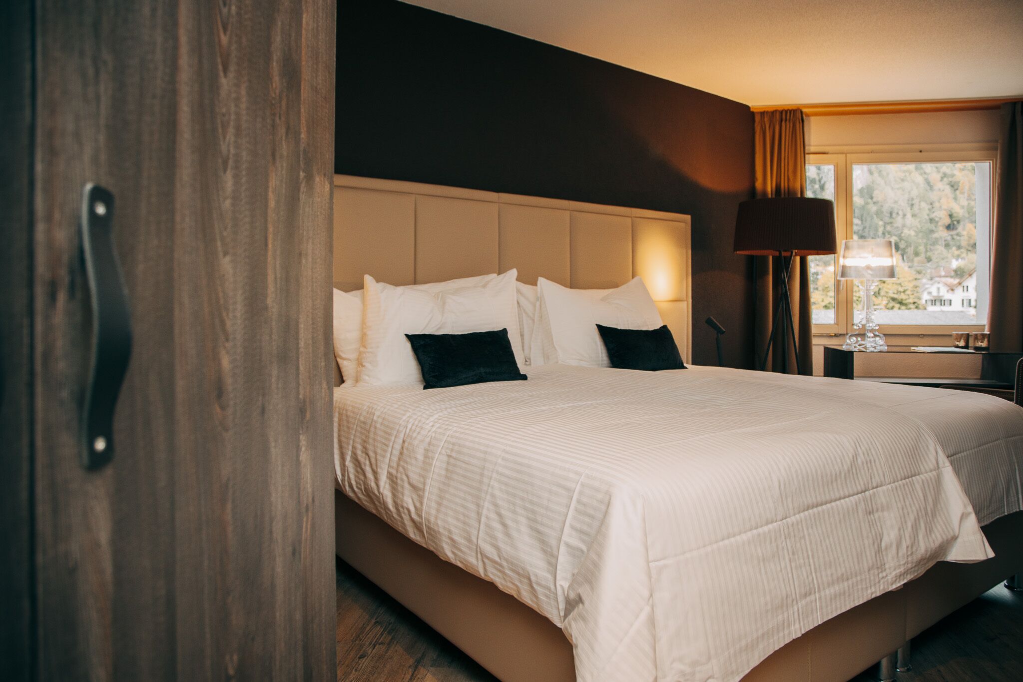 Habitación de hotel con una cama grande, una puerta de madera, una mesita de noche con lámpara y una ventana con vista a árboles.