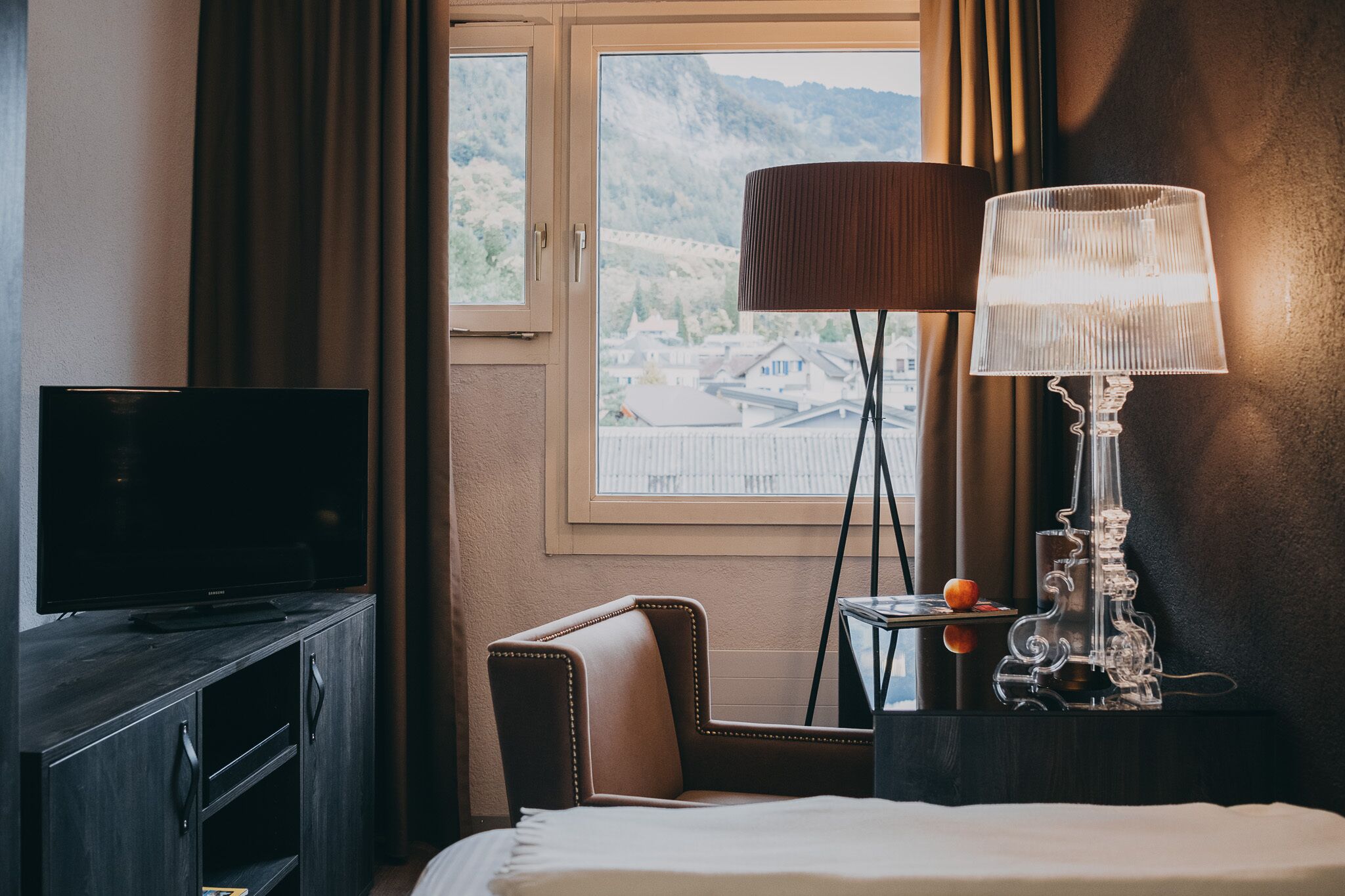 A hotel room with a bed, a television on a wooden cabinet, a chair, a lamp, and a view of a mountainous landscape through a window.
