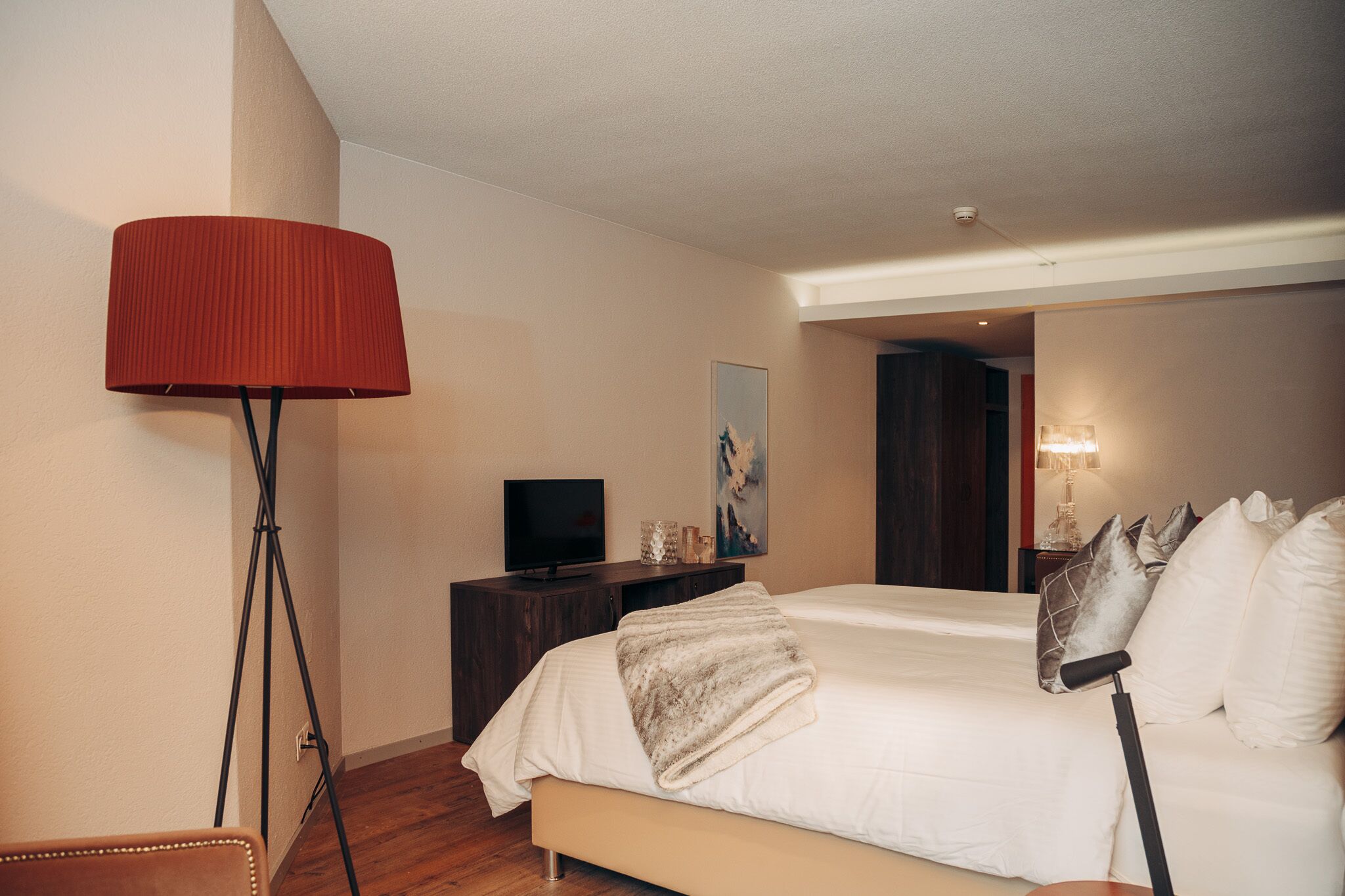Quarto de hotel com uma cama, uma televisão sobre um aparador de madeira, um abajur vermelho e um quadro na parede.