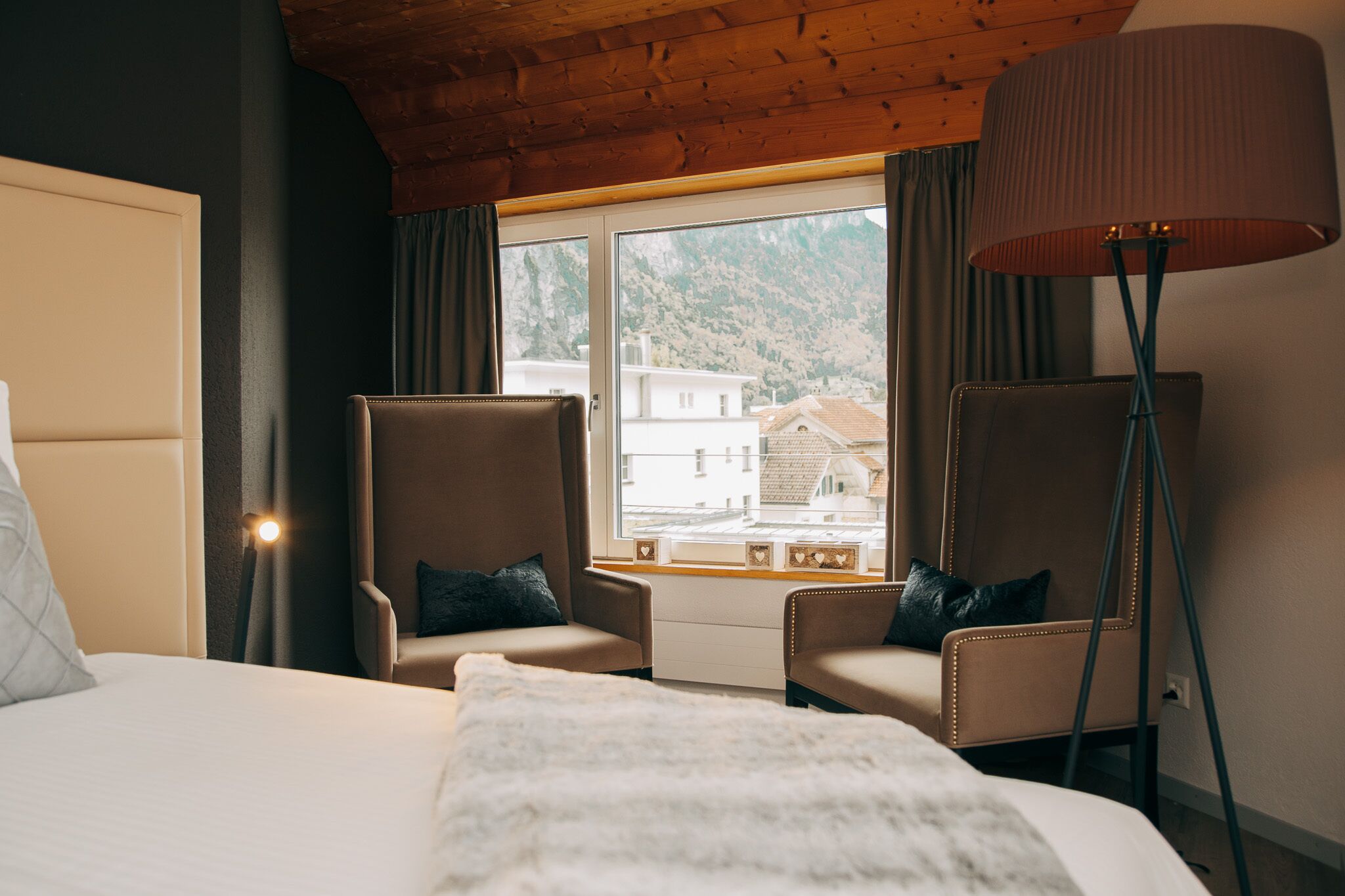 Quarto de hotel com uma cama, dois poltronas, um abajur e uma janela com vista para edifícios e montanhas.