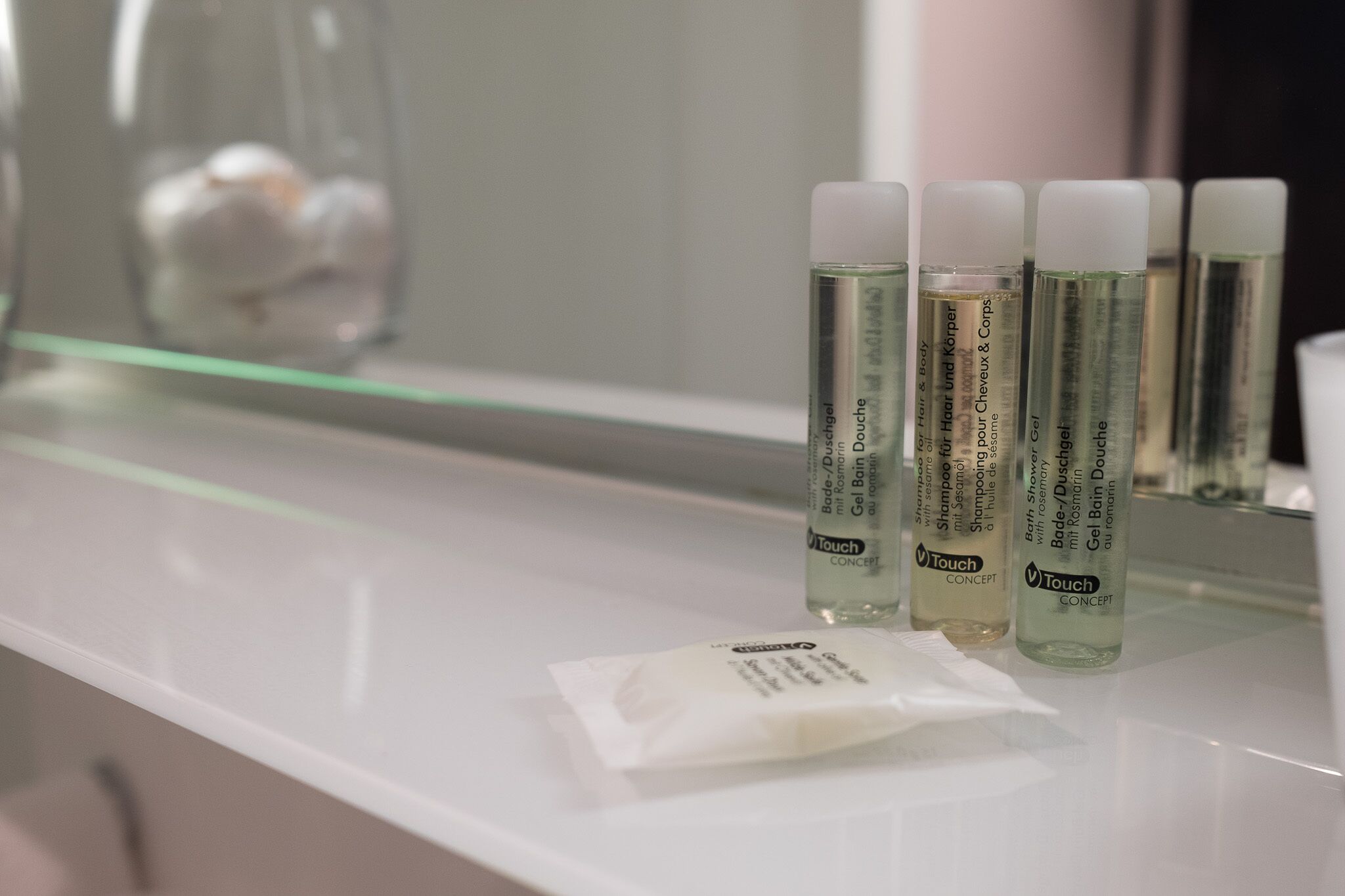 A bathroom counter with three bottles of Touch Concept hair and body care products, a small packet, and a glass jar in the background.