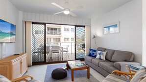 A living room with a sofa, a coffee table, a TV, and a balcony with a view of buildings. - Krait Court, 1 6-8 Krait Cl - great 2 bedroom unit close to little beach (Nelson Bay)