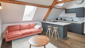 Un espacio tipo loft con sofá, una mesa redonda de madera, zona de cocina con barra y un reloj en la pared.