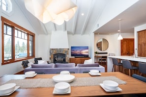 Cozy living area with a dining table for four, fireplace, and TV, offering a view of the outdoors through large windows.