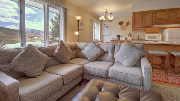 A modern living room with a sectional sofa, a leather ottoman, and a view of a mountainous landscape through large windows. - Cc19 Chateau Claire 2 Bedroom Condo by RedAwning (Dillon)