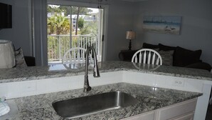 A kitchen with a granite countertop, stainless steel sink, white cabinets, and a view of the outdoors through a large window.