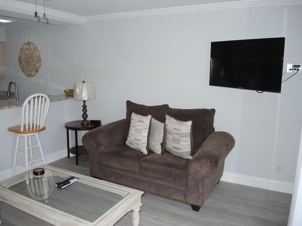 Un salon avec un canapé marron, une table basse en verre, une chaise blanche, une lampe et un téléviseur à écran plat fixé au mur.