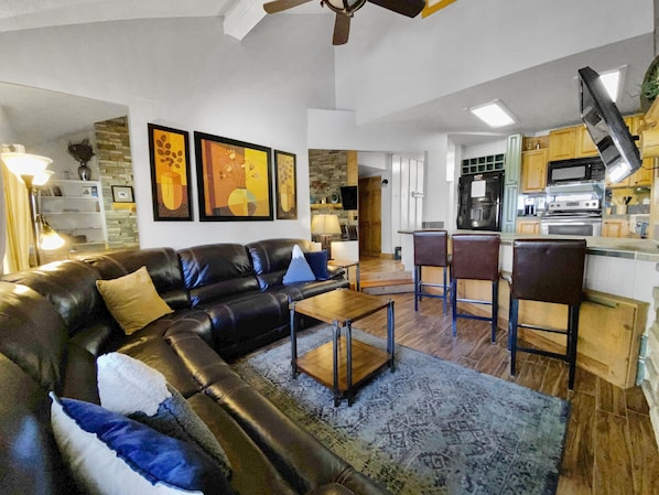 A living room with a leather sofa, coffee table, and patterned rug. There is a kitchen area with bar stools and a refrigerator.